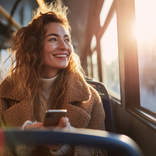 Young woman curly hair brown coat sitting bus holding smartphone sunlight smiling morning city